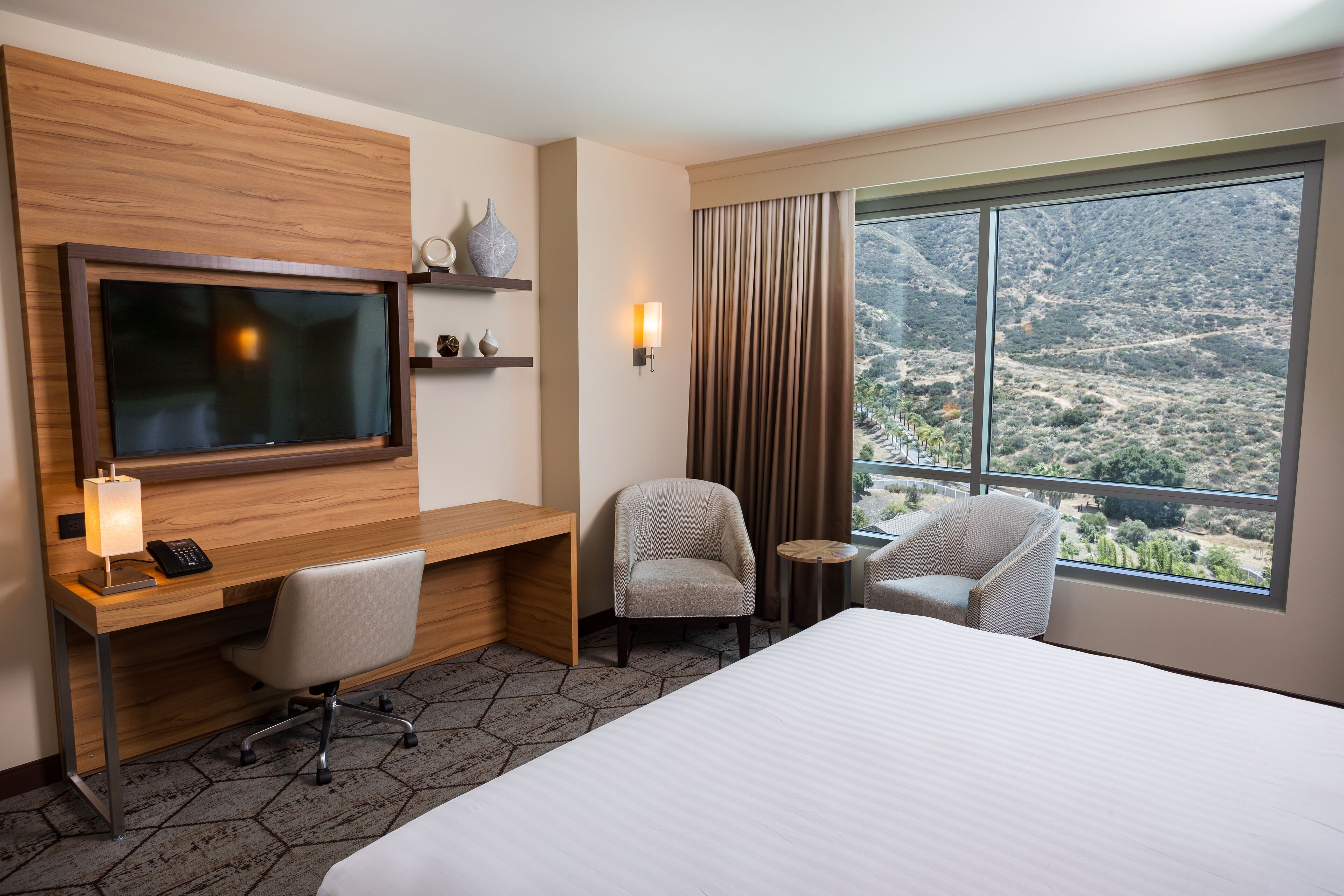 A hotel room with a large bed, a desk with a TV, a chair, a small table, and a view of a mountainous landscape.