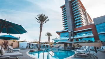 A pool area with lounge chairs and umbrellas, a tall building with balconies, and palm trees.