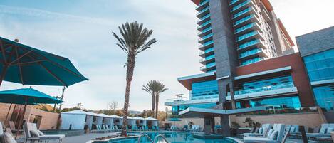 A pool area with lounge chairs and umbrellas, a tall building with balconies, and palm trees.