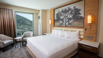 Habitación de hotel con una cama grande, dos sillones, una mesita, una mesita de noche y un cuadro de un árbol.