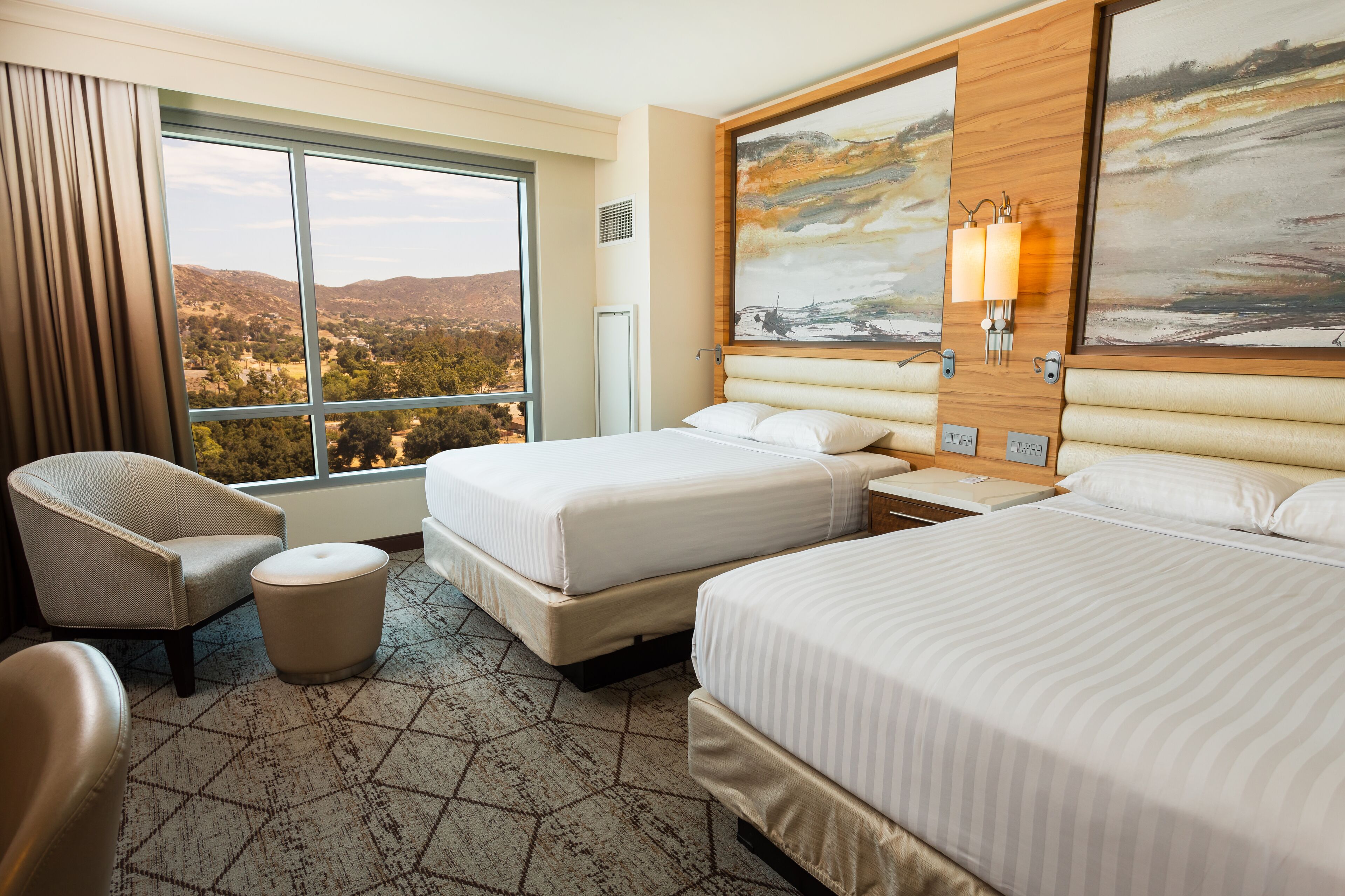A hotel room with two beds, a chair, a small table, and a large window offering a scenic view.