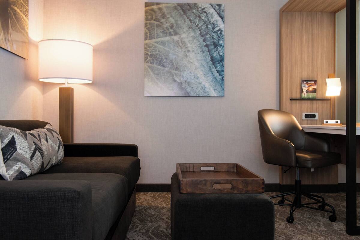 A hotel room with a sofa, a desk, a chair, a lamp, and a painting on the wall.