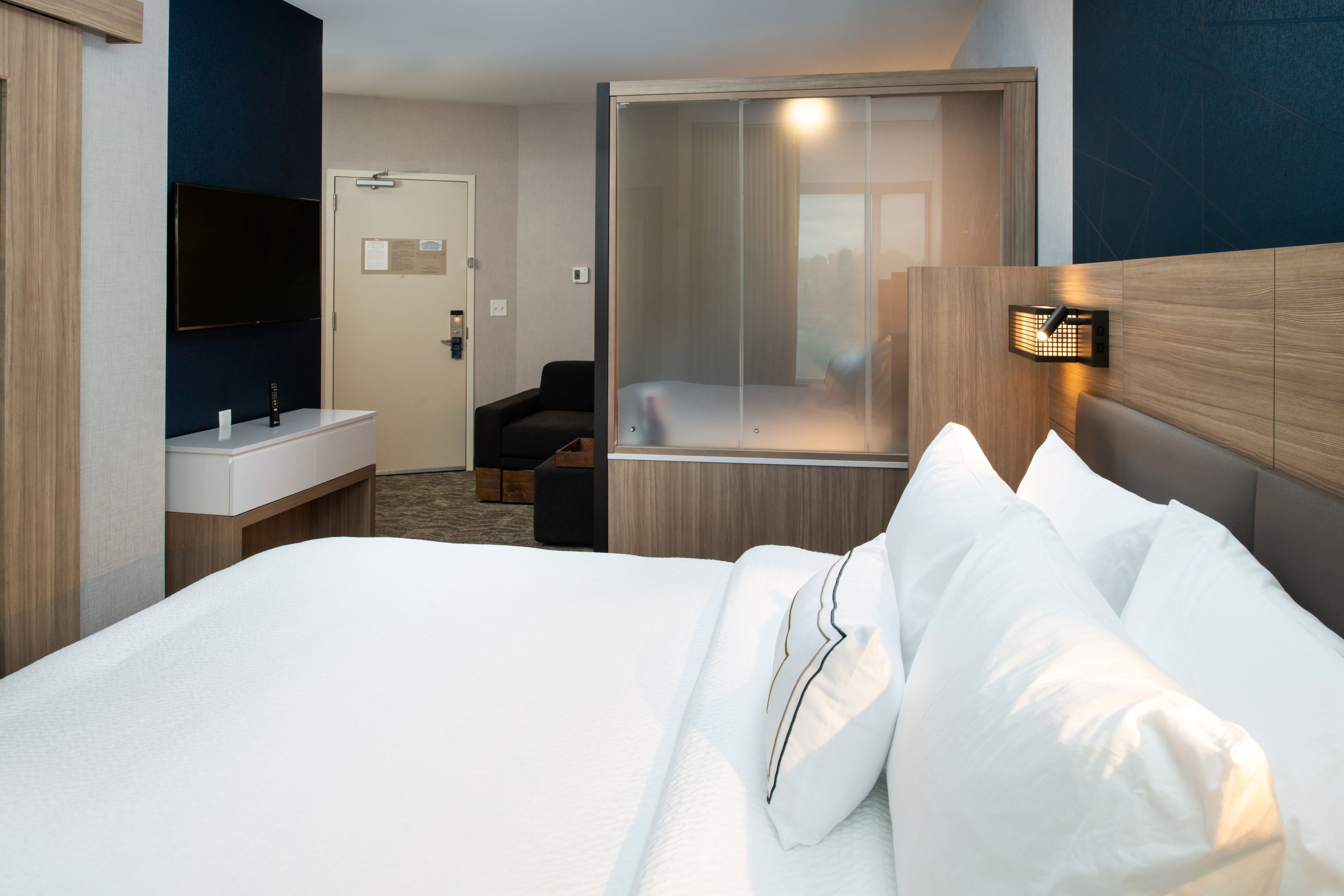 Habitación de hotel con cama, mesita de noche, televisor, cabina de ducha y un pequeño espacio de descanso.