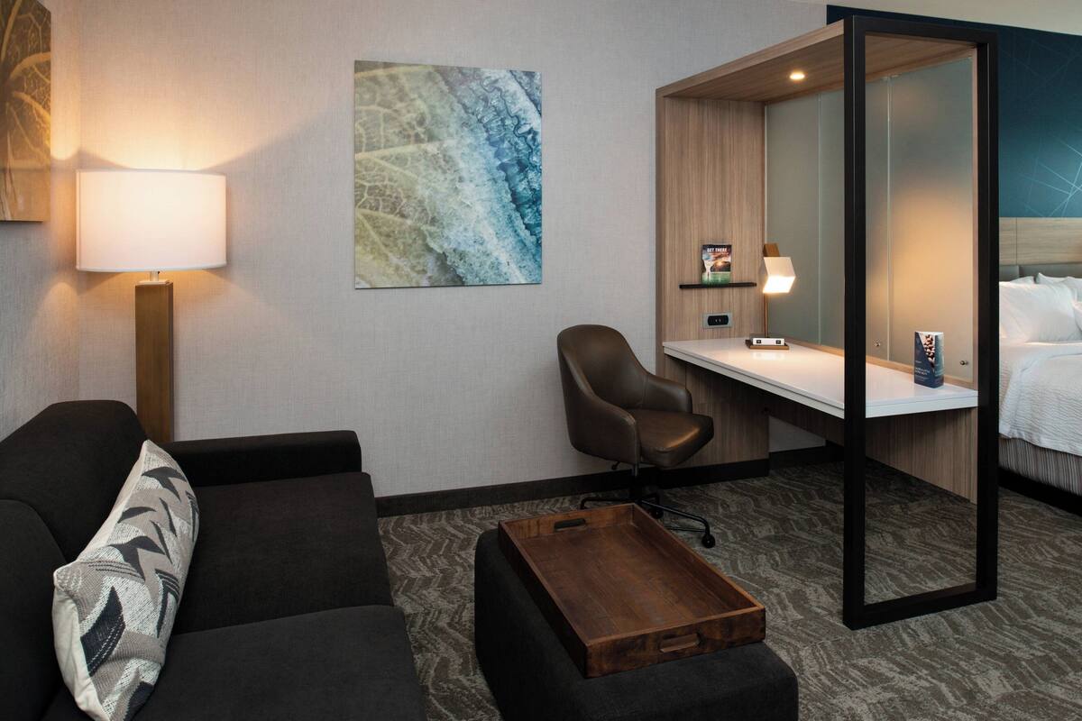 A hotel room with a sofa, a desk, a chair, a bedside table, a lamp, and a bed.
