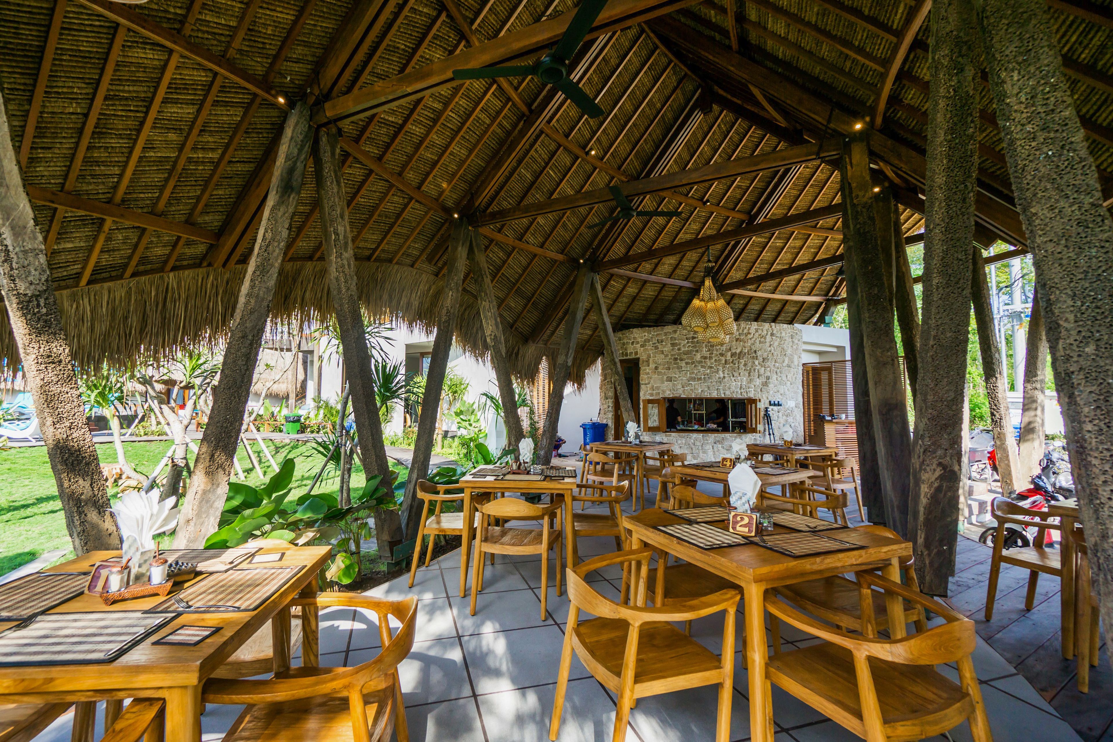 Un restaurante al aire libre con mesas y sillas de madera, techo de paja y pared de piedra.