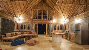 A spacious room with a high ceiling, a stone wall, a wooden bench with cushions, a dining table, and a kitchen area with a refrigerator. - Pondok Manoka (Kintamani)