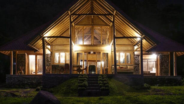 A large, well-lit wooden cabin with a steeply pitched roof, stone foundation, and a covered porch with steps leading to the entrance. - Pondok Manoka (Kintamani)