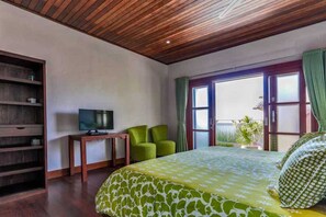 A hotel room with a wooden ceiling, a bed with a green and white patterned bedspread, two green chairs, a television on a wooden table, and a window with green curtains. - Villa Agung (Lembongan Island)