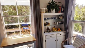 Ein gemütliches Zimmer mit einem Holztisch, zwei Stühlen, einem Korbsessel und einem weißen Schrank mit Regalen, mit Blick auf grüne Pflanzen draußen.
