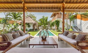 A covered outdoor seating area with two sofas and a table, overlooking a swimming pool and lush garden. - Eco Chic 5BDR Oberoi/ Central Seminyak/beach (Seminyak)