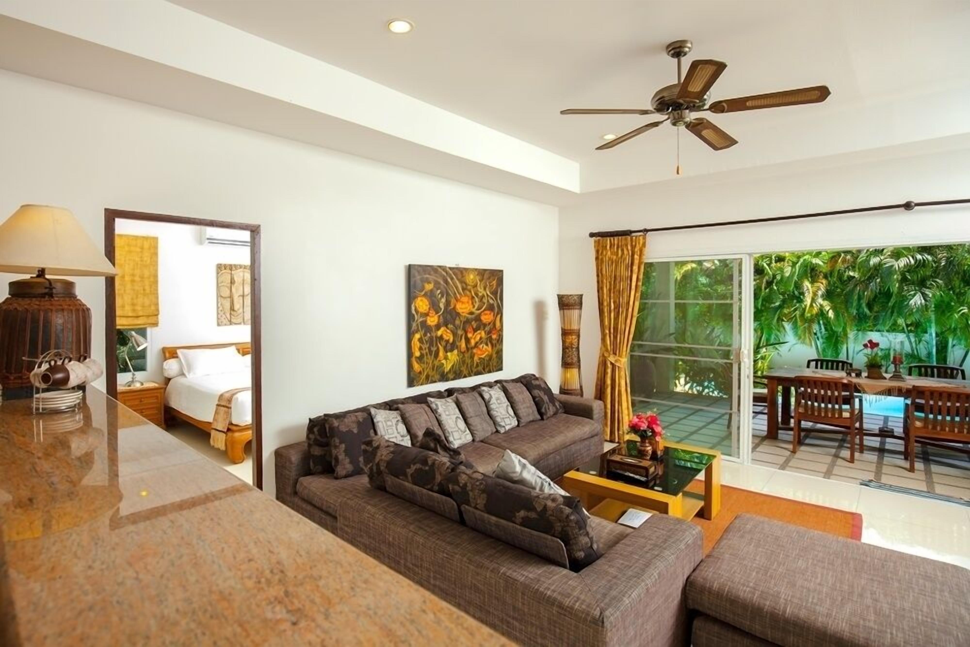 A modern living room with a sofa, coffee table, and a ceiling fan. There is a dining area with a table and chairs visible through a glass door.