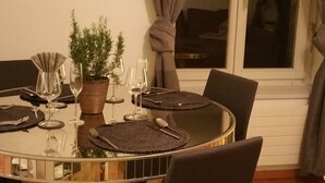 A round dining table with a glass top, surrounded by chairs. There are wine glasses, plates, and cutlery on the table. A potted plant is placed on the table, and a window with curtains is visible in the background. - New:Lovely 2.5 Room , Dish Washer, Wifi, Fully Equiped, own washer/dryer (Zürich)