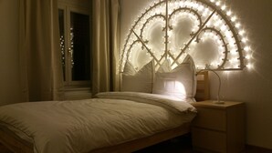 A bedroom with a bed, a nightstand, a window with curtains, and a wall-mounted light fixture. - New:Lovely 2.5 Room , Dish Washer, Wifi, Fully Equiped, own washer/dryer (Zürich)