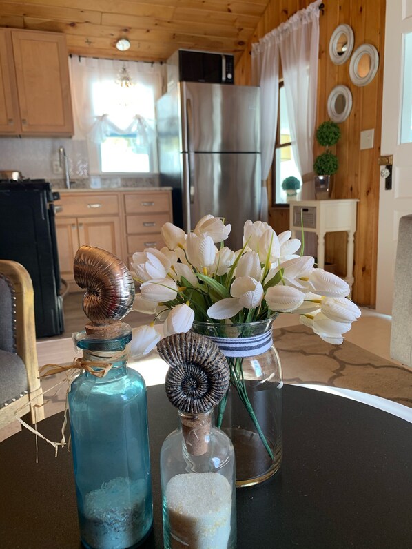 Una cocina con armarios de madera, un refrigerador de acero inoxidable y una zona de comedor con una mesa que presenta botellas decorativas y un florero con flores blancas.