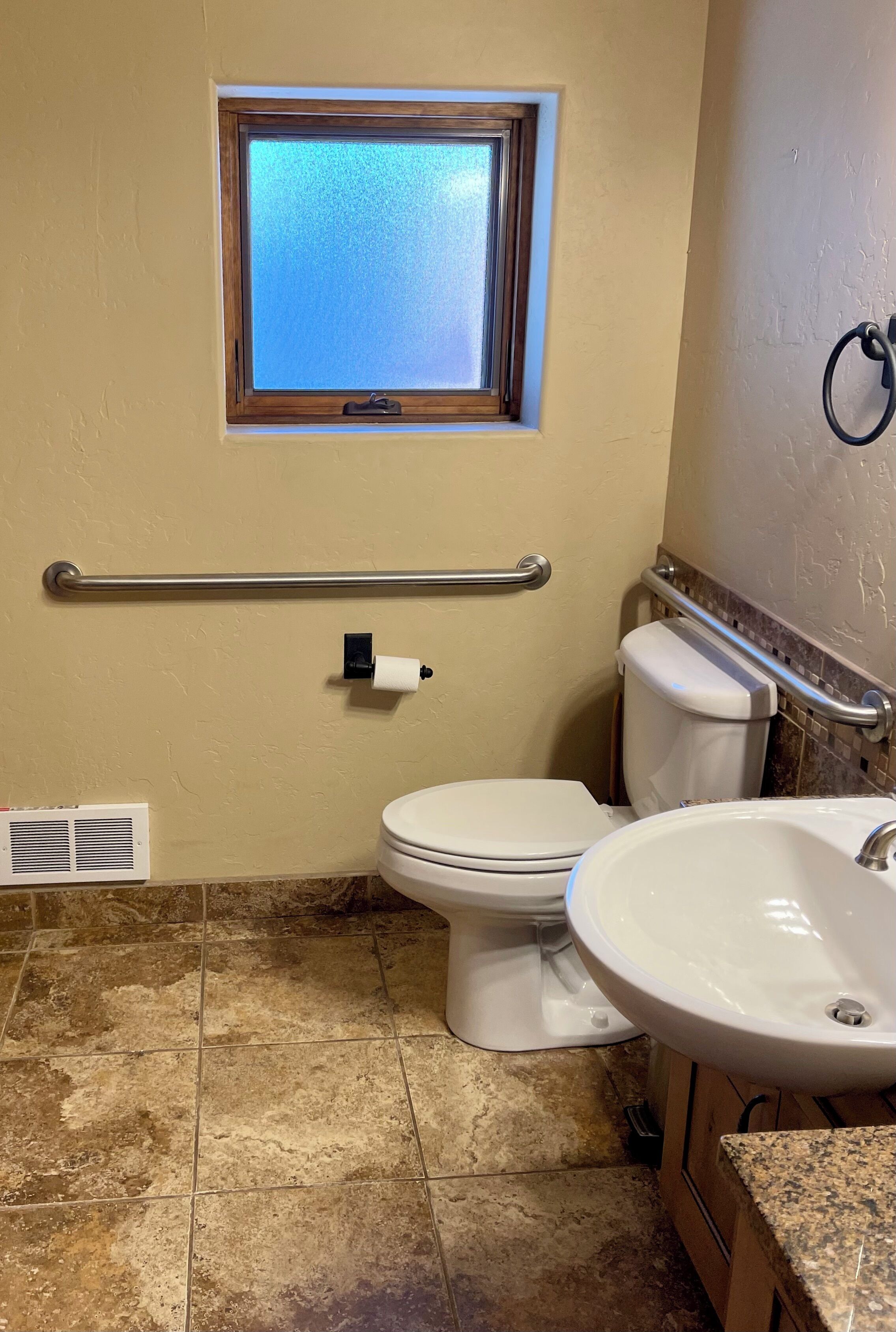 Une salle de bain équipée de toilettes, d’un lavabo, d’une fenêtre et d’une barre d’appui.