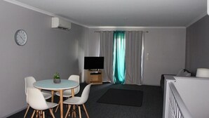A modern, compact living space with a round dining table, white chairs, a flat-screen TV, a small plant, and a window with curtains.