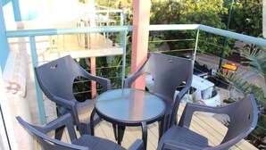 A balcony with a glass table and four chairs, surrounded by a railing and greenery.