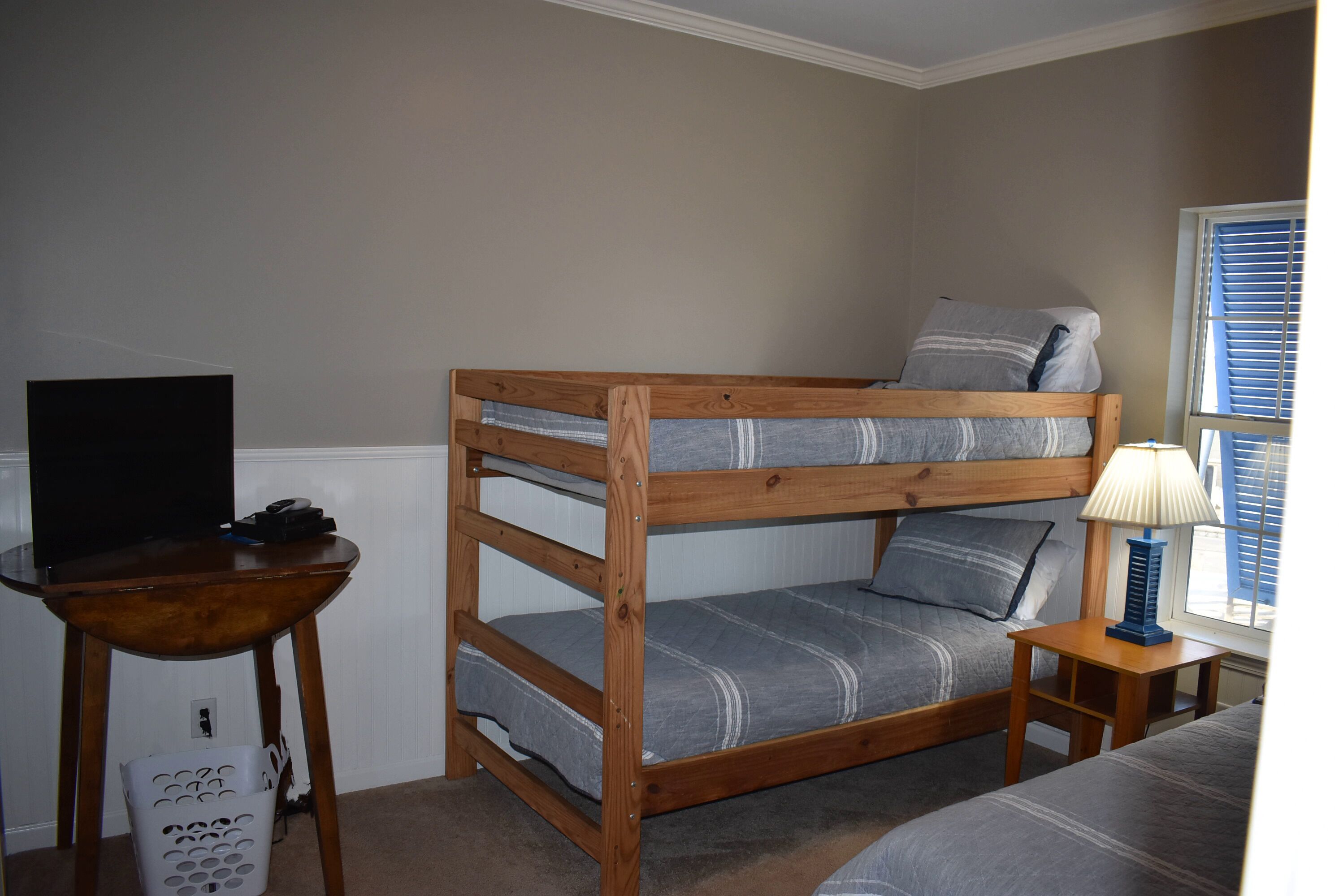 A bunk bed with a TV on a wooden table beside it, a lamp on a small table, and a laundry basket.