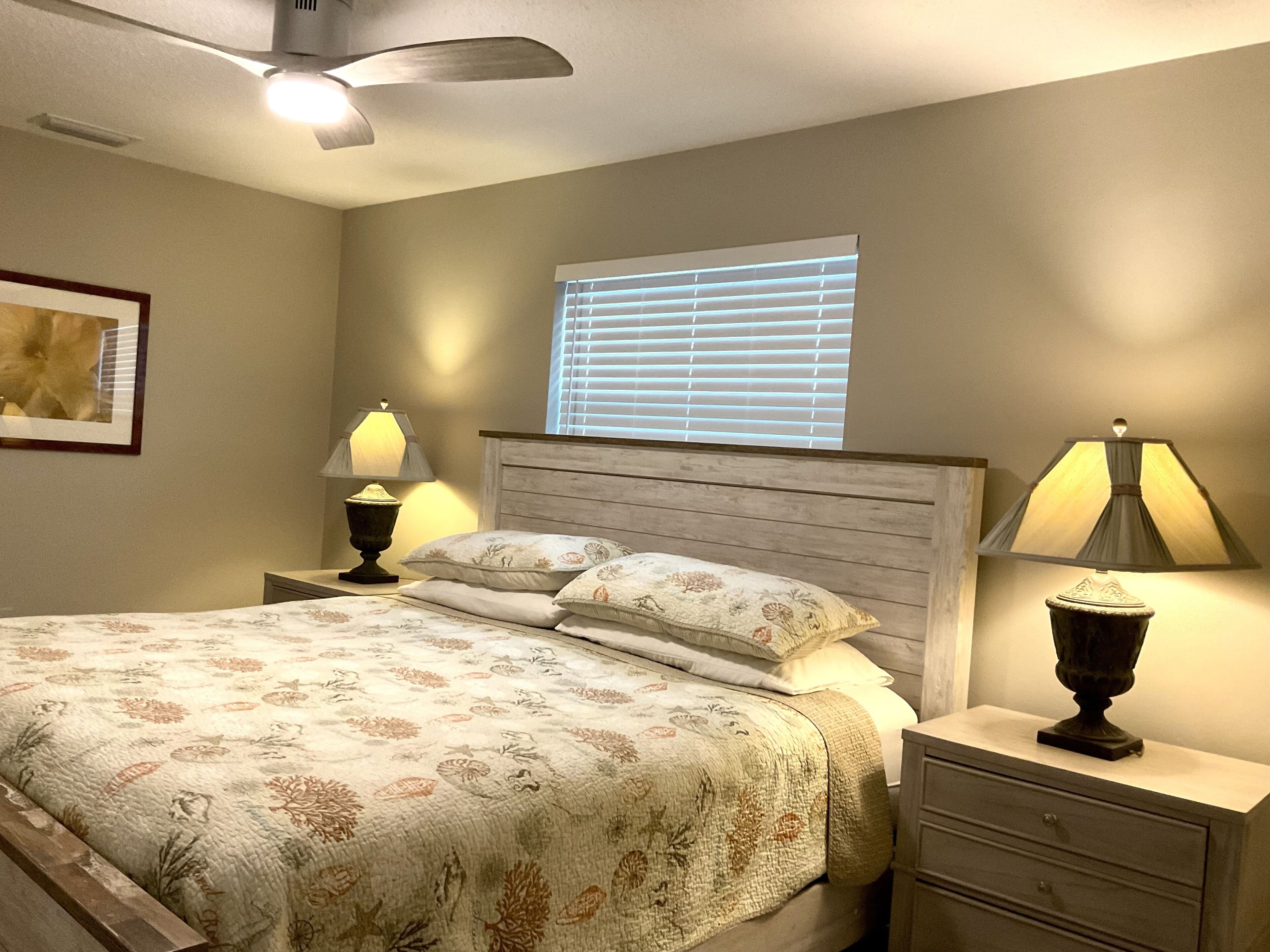 Ein Schlafzimmer mit einem Bett, zwei Nachttischen mit Lampen, einem Deckenventilator, einem Fenster mit Jalousien und einem gerahmten Bild an der Wand.