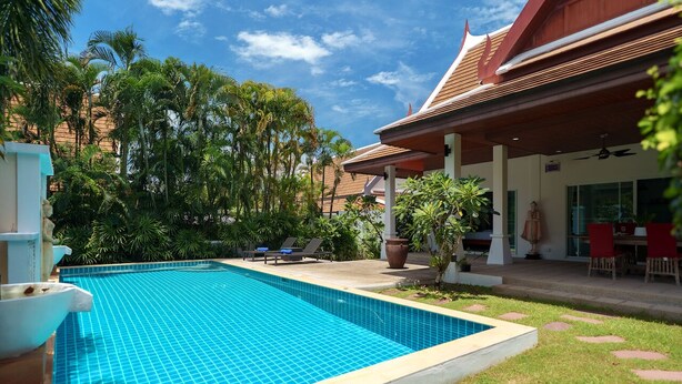 A swimming pool with tiled edges, surrounded by a grassy area, palm trees, and a covered patio with outdoor seating.