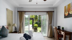 A hotel room with a bed, a desk, a chair, and a view of a garden through large windows.