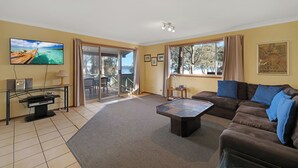 A living room with a large TV, a sofa, a coffee table, and a view of the outdoors. - Absolute Waterfront - large deck, level access to water, plenty wildlife (St Georges Basin)