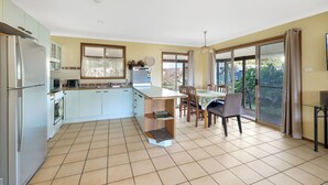 A modern kitchen with a central island, dining area with chairs, and large sliding glass doors to a balcony. - Absolute Waterfront - large deck, level access to water, plenty wildlife (St Georges Basin)