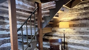 Un intérieur rustique avec un escalier en bois, une chaise en bois recouverte d’une peau de bête et une lampe.