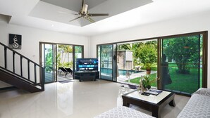 Amplia sala de estar con televisor grande, ventilador de techo y vista a un jardín a través de puertas correderas.