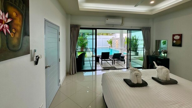 Ein Hotelzimmer mit einem großen Bett, einem Deckenventilator, einer Schiebetür zu einem Balkon mit Sitzgelegenheit und Blick auf einen Swimmingpool.