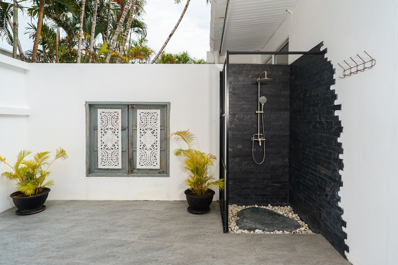 Un área de ducha exterior moderna con un muro de azulejos negros, dos plantas en macetas y una ventana decorativa con cortinas de encaje.