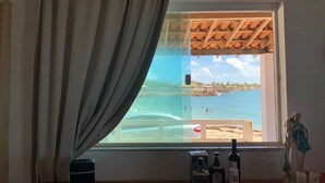Une chambre avec vue sur une plage et l’océan, une fenêtre avec un rideau et une statue posée sur le rebord de la fenêtre.