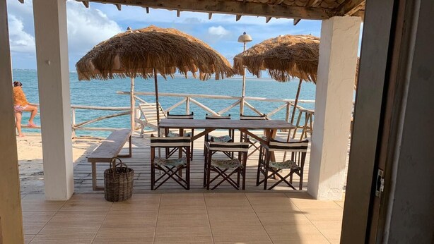 Une terrasse au bord de la plage, avec des tables et des chaises en bois, des parasols en chaume et une vue sur l’océan.