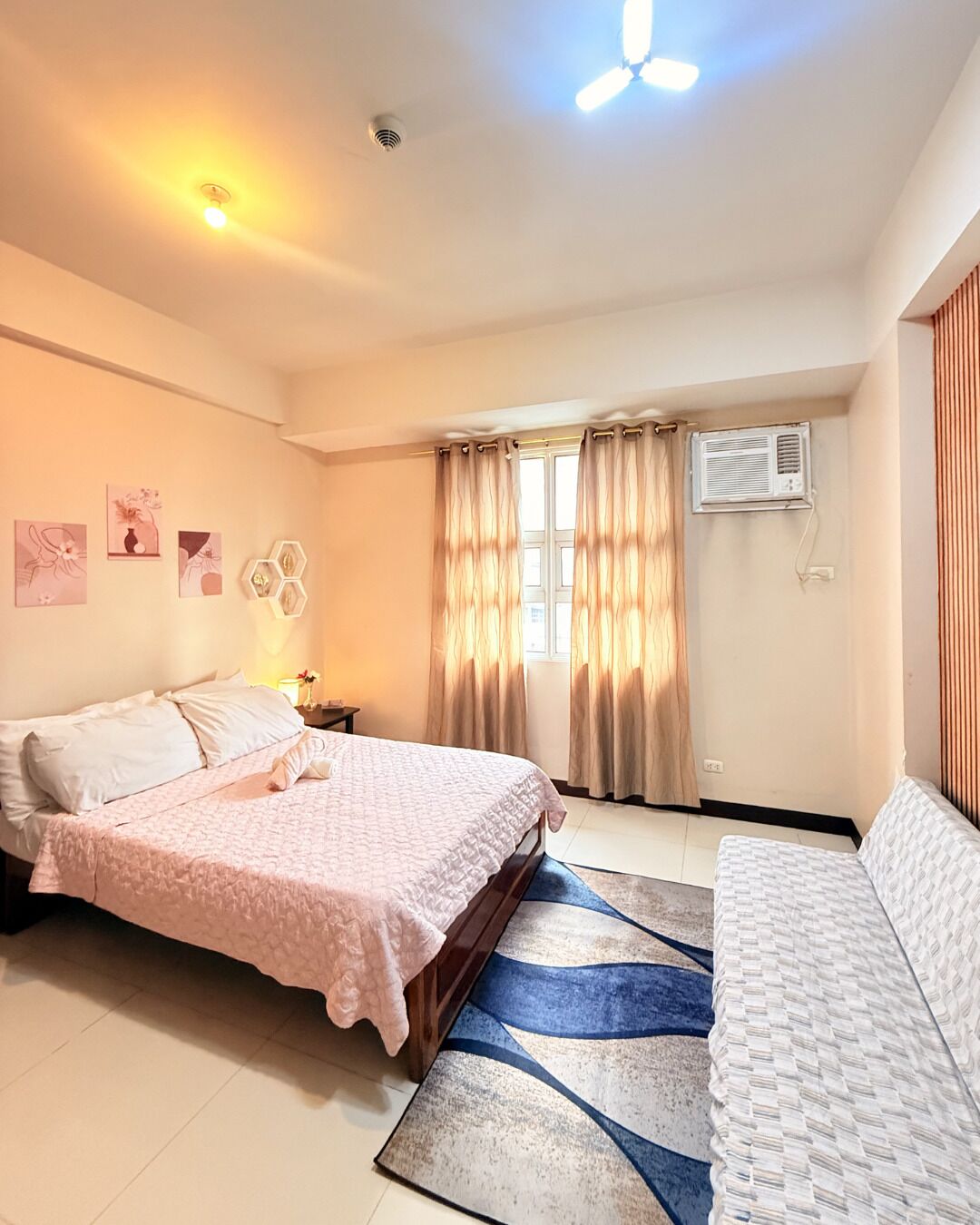 A bedroom with a bed, a sofa, a window with curtains, an air conditioning unit, and wall art.