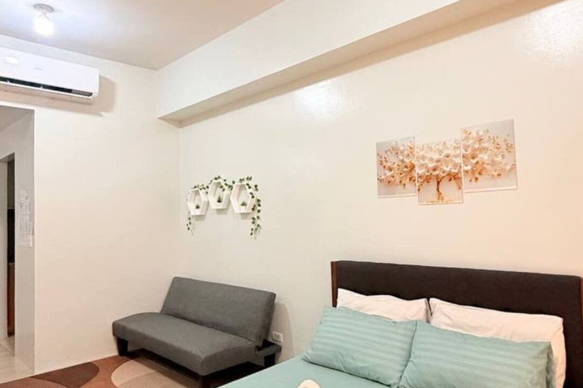 Un dormitorio con cama, sofá, aire acondicionado instalado en la pared y una pequeña planta decorativa.