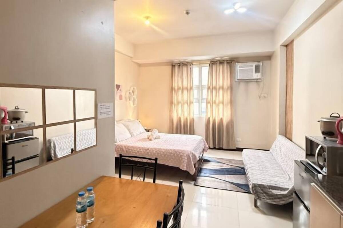 Una habitación de hotel compacta con cama, sofá, mesa de comedor con sillas, kitchenette y ventana con cortinas.