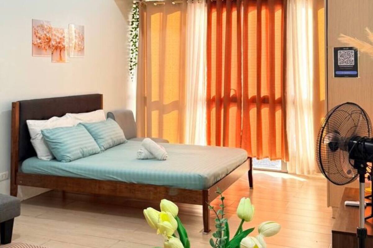 Un dormitorio con cama, una mesita de noche con un florero, un ventilador de pie y una ventana con cortinas.