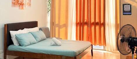 A bedroom with a bed, a bedside table with a vase of flowers, a standing fan, and a window with curtains.
