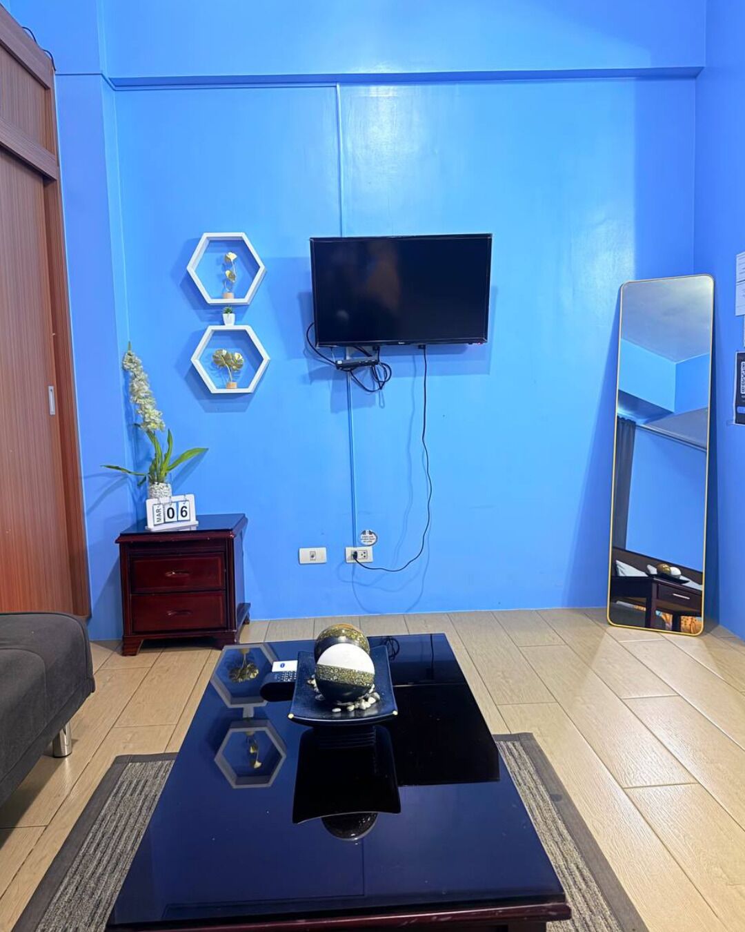 A room with a blue wall, a mounted TV, a wooden cabinet, a small table with decorative items, and a mirror.