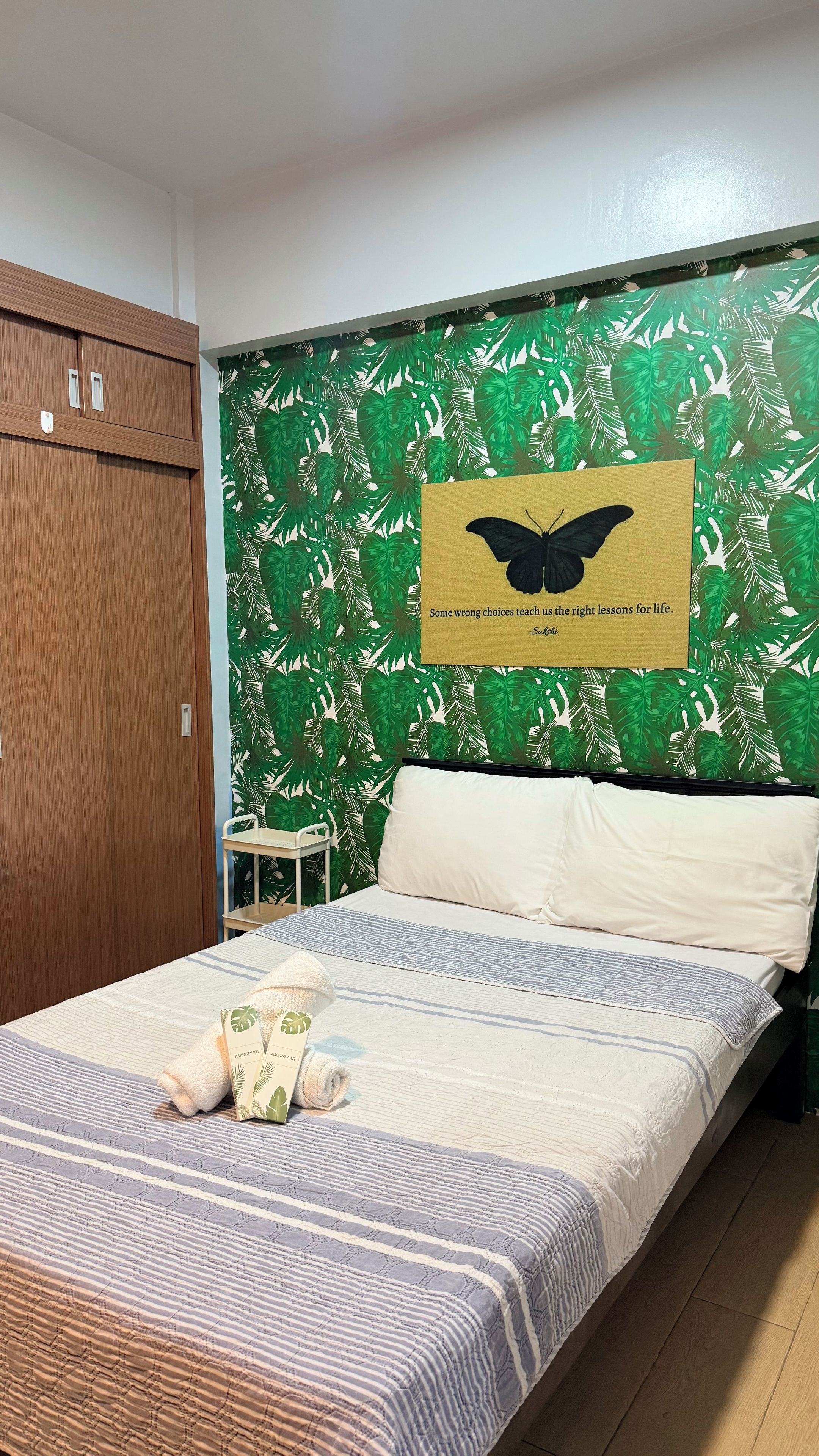 Un dormitorio con cama, armario de madera, una mesita y pared con diseño de hojas.