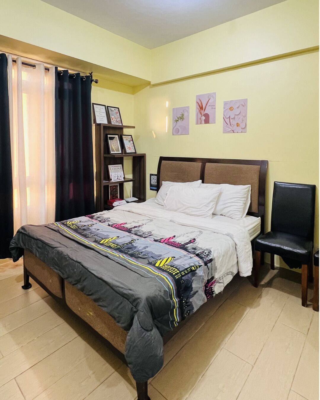 A bedroom with a bed, a chair, a shelf with framed photos, and a wall with floral artwork.