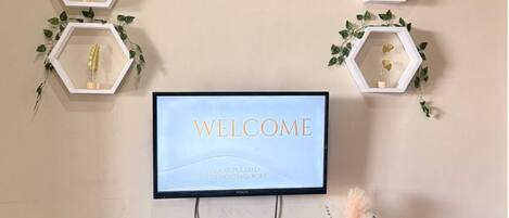 A wall-mounted clock, a welcome message on a TV, and decorative hexagonal frames with plants.