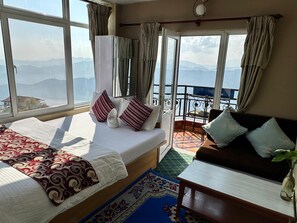 A hotel room with a bed, a sofa, a small table, and a balcony with a view of mountains.