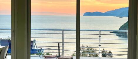 A room with a large sliding glass door leading to a balcony with a view of the sea and distant mountains.