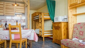A compact kitchen with wooden cabinets, a dining table with chairs, a bunk bed, and a microwave on a wooden cabinet. - Cosy apartment for 4 guests with WIFI, TV and balcony (Les Boisses)