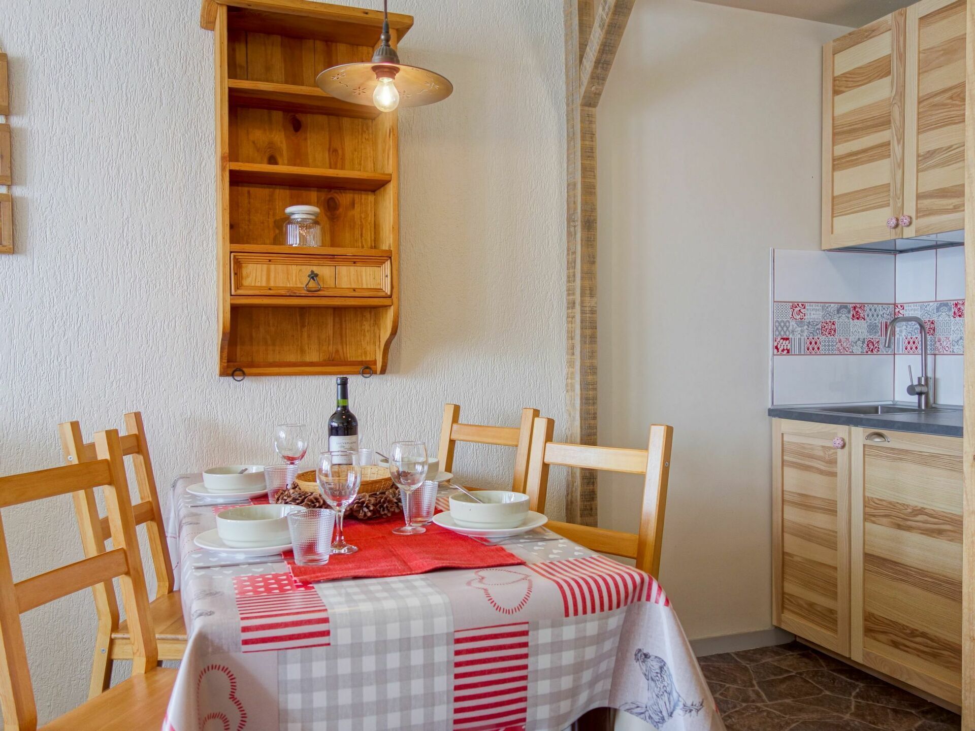 A dining area with a table set for four, wooden chairs, a red and white checkered tablecloth, a hanging light fixture, and a kitchen with wooden cabinets and patterned backsplash tiles.