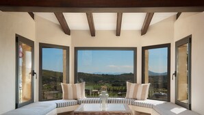 Une pièce spacieuse dotée d’une grande fenêtre donnant sur un paysage verdoyant. On y trouve une table basse en bois et deux coins salon confortables, avec des coussins à rayures.