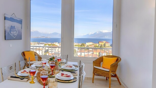 Ein Essbereich mit Tisch für zwei Personen, zwei Korbstühle und Blick auf eine Küstenstadt sowie Berge durch große Fenster.