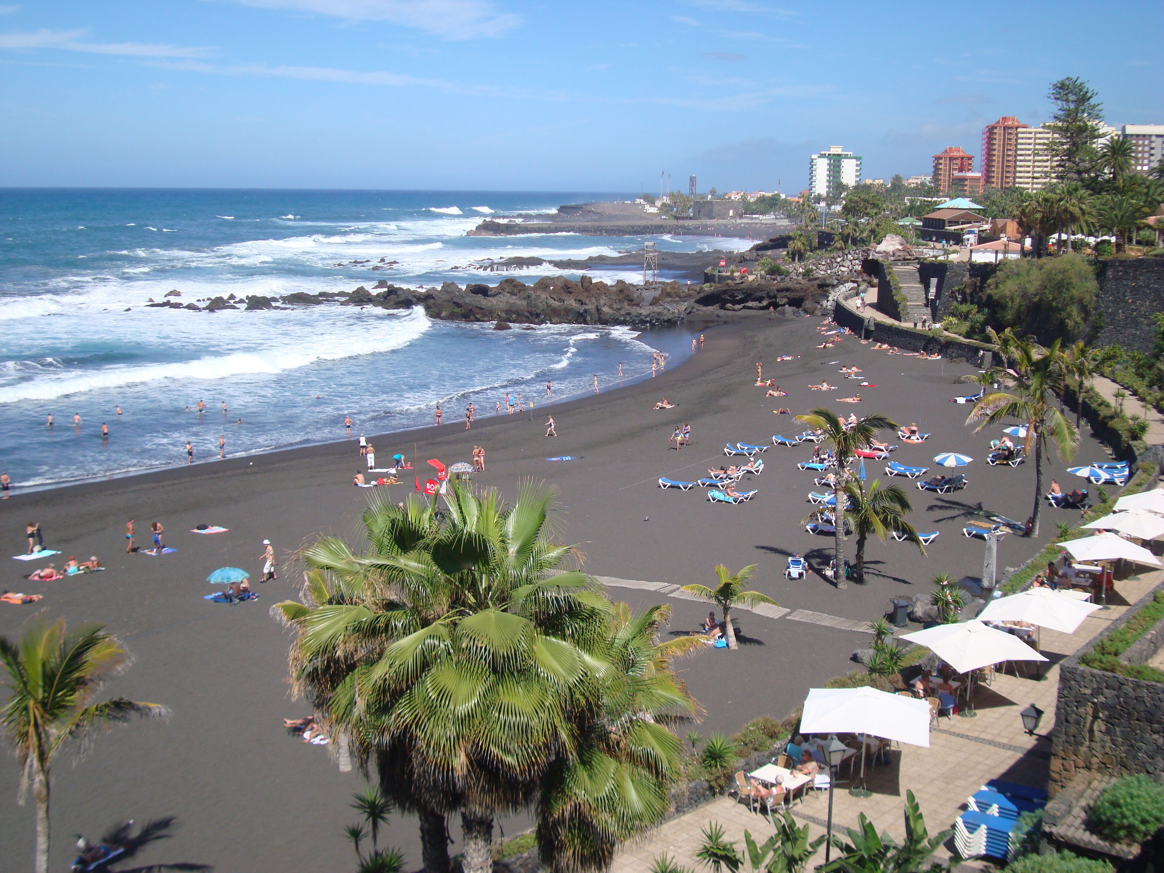 Una playa con sombrillas y sillas reclinables, un litoral rocoso y edificios al fondo.