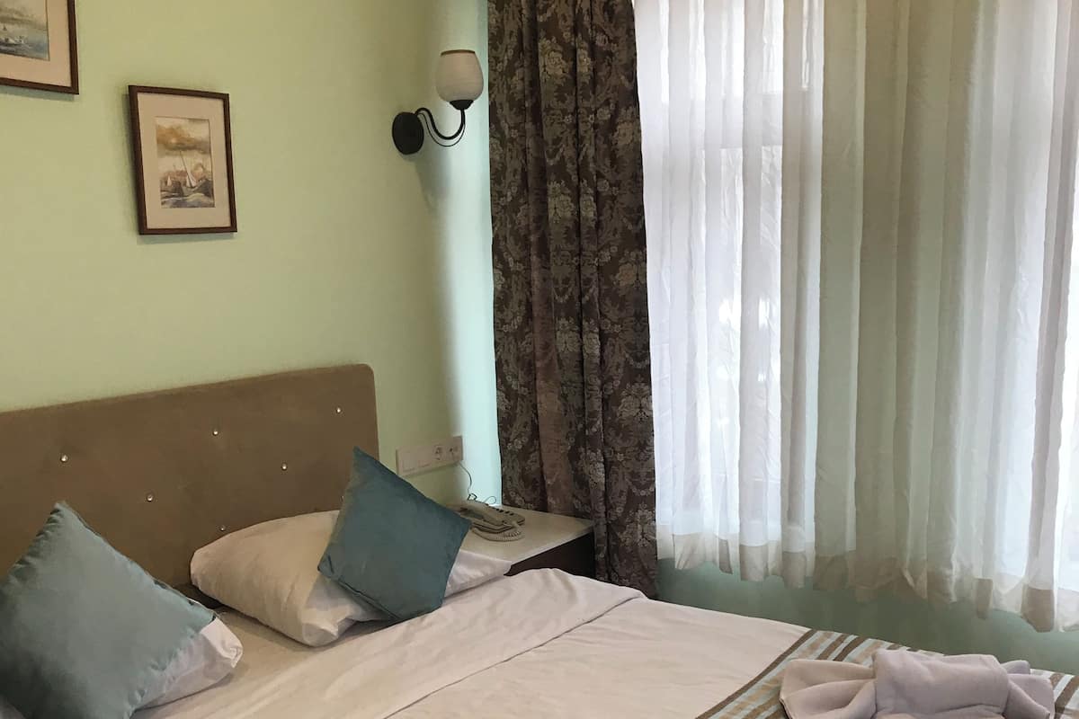 A neatly made bed with pillows, a striped blanket, and towels on the bed. There is a nightstand with a lamp and a phone. The room has light green walls, a window with curtains, and framed pictures on the wall.